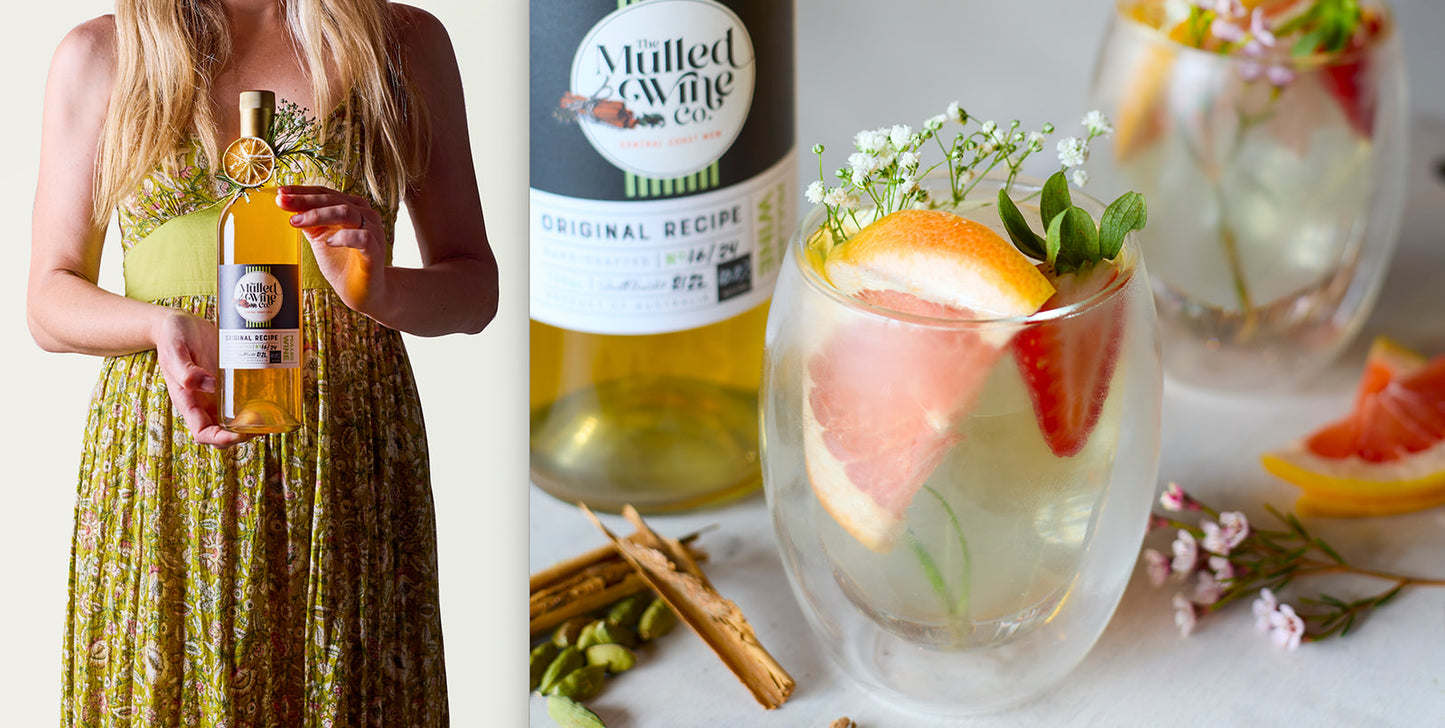 Women in a summery dress and long blonde hair holding a beautifully decorated bottle of white mulled wine along side double-walled glasses filled with wine and fruit.