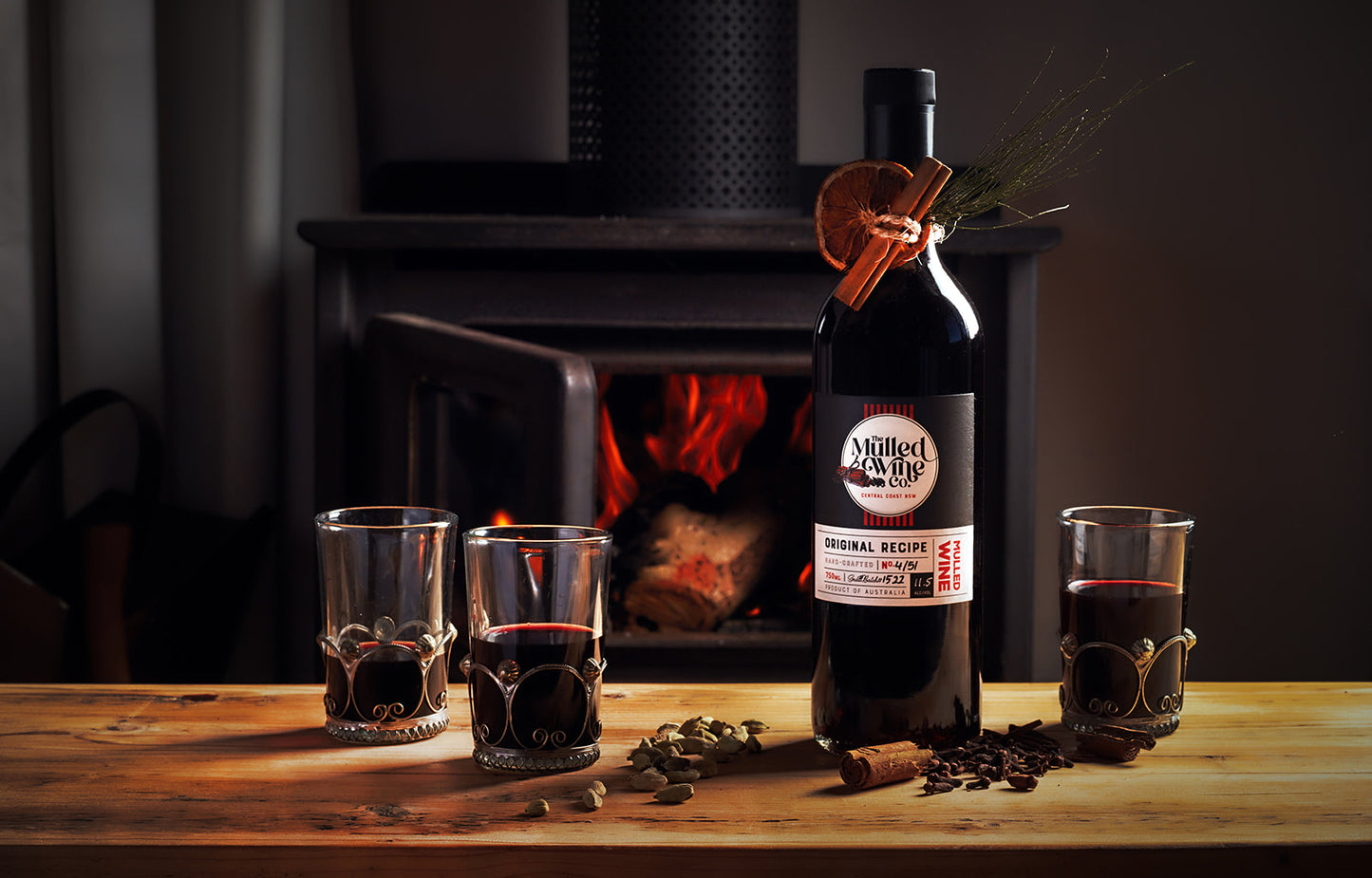 Glasses and a decorated bottle of Original Recipe red mulled wine in front of a fireplace.